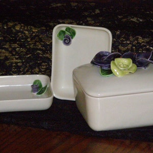 May include: Three white ceramic boxes with floral designs. The largest box has a lid and a purple and green flower design. The medium box has a green flower design. The smallest box is rectangular and has a purple flower design.