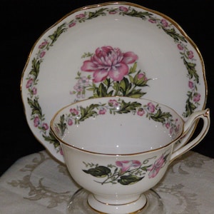 May include: A white teacup and saucer with a gold rim and floral pattern. The saucer features a pink flower with green leaves and a border of pink and green flowers. The teacup has a similar floral pattern with a pink flower and green leaves.