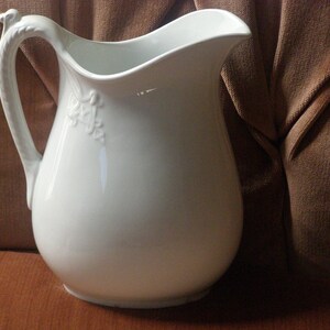 May include: A white ceramic pitcher with a decorative floral design on the side. The pitcher has a wide mouth and a handle that curves up and over the top.