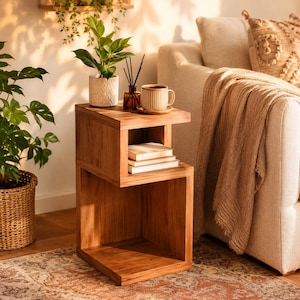 May include: A wooden side table with an S-shaped profile featuring an open top surface and a lower storage compartment. The table holds a small potted plant, a glass reed diffuser, and a ribbed ceramic mug, with two books stacked in the middle shelf.