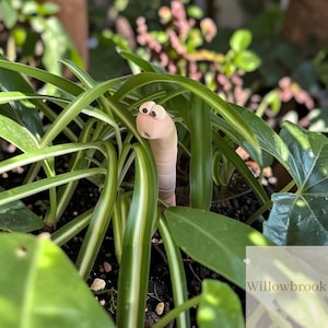 Puede incluir: Una figura de gusano rosa claro con ojos de dibujos animados emerge de una planta verde con hojas largas y rayadas. El gusano está posicionado entre el follaje, creando una escena de jardín lúdica. La planta tiene rayas blancas.