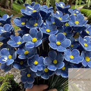 May include: A bunch of blue ceramic flowers with yellow centres. The flowers have a glossy finish and are arranged closely together. The stems are green, and the background is blurred, suggesting an outdoor setting. The text "Willowbrook" is visible in the bottom right corner.