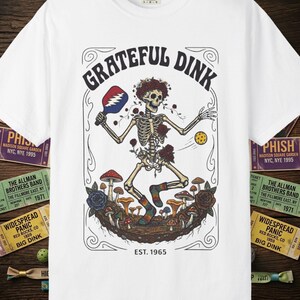 May include: White t-shirt with a graphic featuring a skeleton playing pickleball, surrounded by mushrooms and roses. The text "GRATEFUL DINK" arches above the skeleton, with "EST. 1965" below. Concert tickets are visible on the sides.