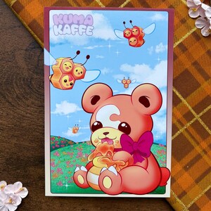 May include: A rectangular art print featuring the text KUMA KAFFE. The illustration depicts a coral-colored bear with a white crescent mark on its forehead, wearing a dark pink bow, eating honey from a pot. Three small bee-like creatures fly above.