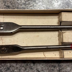 May include: A set of two metal expansive drill bits with adjustable cutters and threaded pilot tips, housed in a rectangular cardboard box. The bits feature silver-toned shafts with red-painted ends and include two additional loose metal cutters.