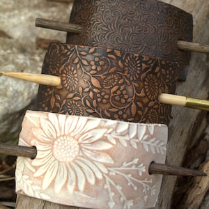 May include: Three leather hair sticks with floral patterns. The top stick has a brown floral pattern, the middle stick has a brown and copper floral pattern, and the bottom stick has a white floral pattern on a light brown background.