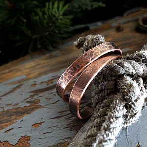 May include: Two copper bracelets with a hammered texture rest on a thick, gray rope. The bracelets are a warm, reddish-brown color, and the rope is knotted. The background includes weathered wood and evergreen branches.