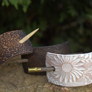 May include: Three leather hair clips with different floral designs. The clips are brown, dark brown, and white. Each clip has a wooden hair stick.