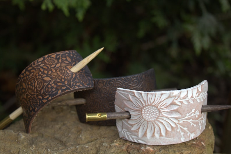 May include: Three leather hair clips with different floral designs. The clips are brown, dark brown, and white. Each clip has a wooden hair stick.