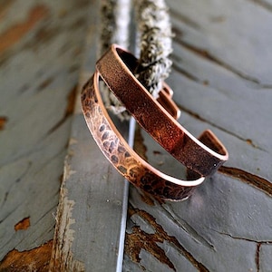 May include: Two copper cuff bracelets with textured surfaces. One bracelet has a hammered pattern, while the other has a smooth finish. The bracelets are displayed on a weathered wooden surface, showcasing their rustic charm. Jewelry.