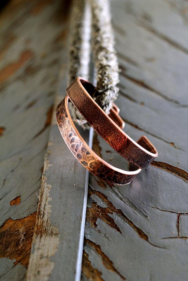 May include: Two copper cuff bracelets with textured surfaces. One bracelet has a hammered pattern, while the other has a smooth finish. The bracelets are displayed on a weathered wooden surface, showcasing their rustic charm. Jewelry.