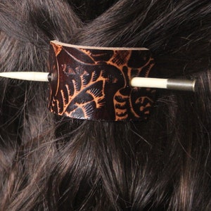 Puede incluir: Una pinza para el pelo de cuero marr&oacute;n oscuro con un intrincado dise&ntilde;o tallado. El accesorio para el cabello presenta una forma cil&iacute;ndrica con un palito de madera de color claro y un acento de metal plateado. La pinza est&aacute; insertada en cabello casta&ntilde;o oscuro.