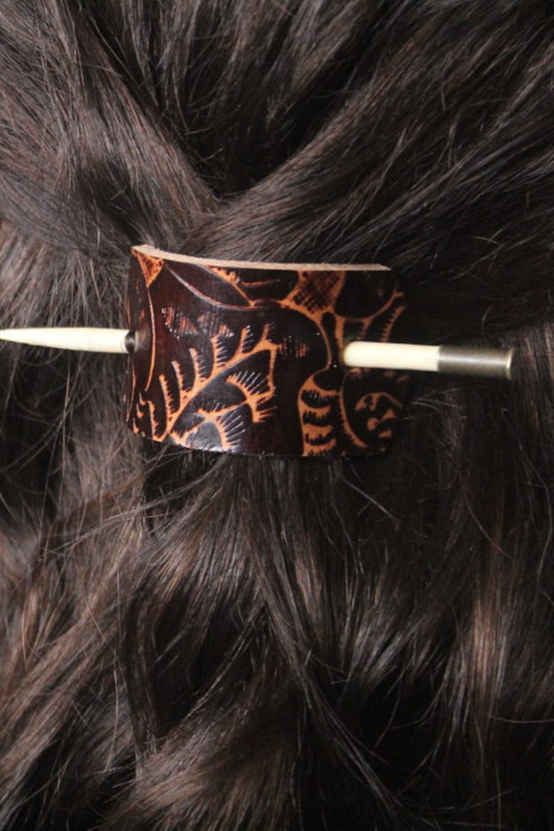 Puede incluir: Una pinza para el pelo de cuero marr&oacute;n oscuro con un intrincado dise&ntilde;o tallado. El accesorio para el cabello presenta una forma cil&iacute;ndrica con un palito de madera de color claro y un acento de metal plateado. La pinza est&aacute; insertada en cabello casta&ntilde;o oscuro.