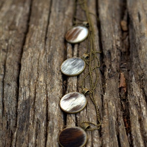 May include: A necklace with four round pendants made of cowhide with a brown and white pattern. The necklace is made of a brass chain and is displayed on a piece of weathered wood.