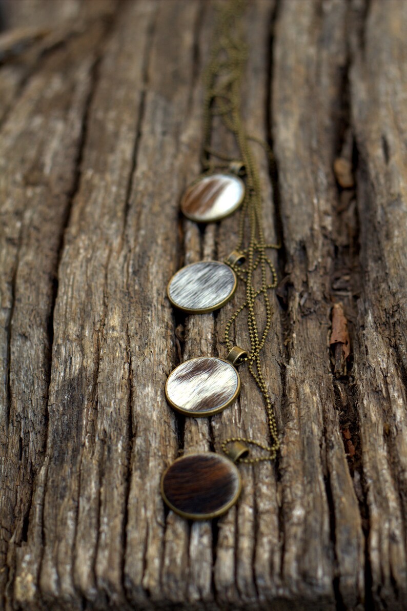 May include: A necklace with four round pendants made of cowhide with a brown and white pattern. The necklace is made of a brass chain and is displayed on a piece of weathered wood.