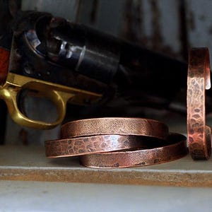 May include: Copper bracelets with a hammered texture are displayed alongside a vintage revolver with a wooden grip and brass trigger. The bracelets are stacked and stand upright, showcasing their rustic design.