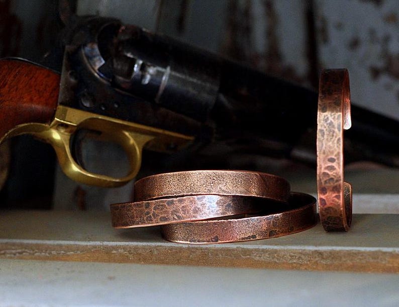 May include: Copper bracelets with a hammered texture are displayed alongside a vintage revolver with a wooden grip and brass trigger. The bracelets are stacked and stand upright, showcasing their rustic design.