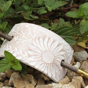 May include: A white ceramic hair clip with a floral design and a wooden stick. The clip is shaped like a half-circle and has a raised sunflower design. The stick is brown and has a gold-colored tip.