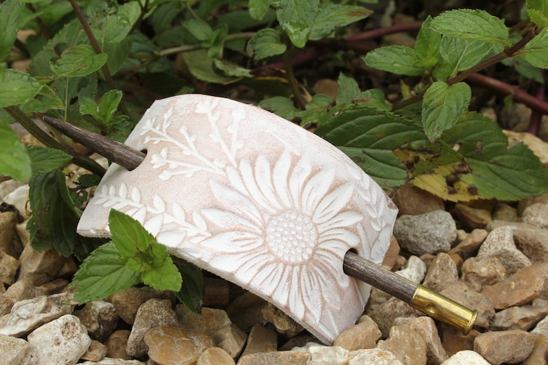 May include: A white ceramic hair clip with a floral design and a wooden stick. The clip is shaped like a half-circle and has a raised sunflower design. The stick is brown and has a gold-colored tip.