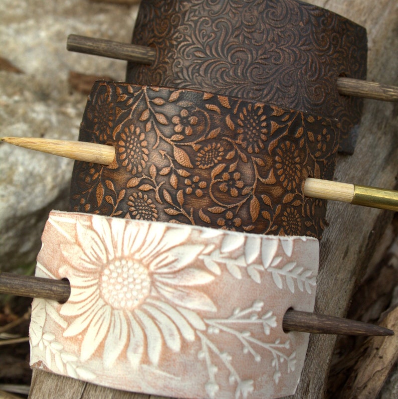 May include: Three leather hair sticks with floral patterns. The top stick has a brown floral pattern, the middle stick has a brown and copper floral pattern, and the bottom stick has a white floral pattern on a light brown background.