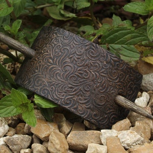 May include: A brown leather hair stick with a decorative floral pattern. The stick is made of wood and has a dark brown finish.