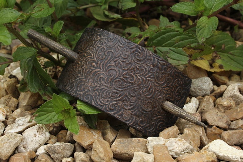 May include: A brown leather hair stick with a decorative floral pattern. The stick is made of wood and has a dark brown finish.