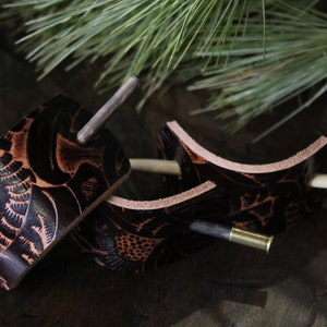 Puede incluir: Tres horquillas artesanales de cuero con intrincados dise&ntilde;os florales. El cuero marr&oacute;n oscuro se acent&uacute;a con detalles en marr&oacute;n claro. Cada horquilla presenta un pasador de metal para asegurar el cabello. Un accesorio natural y r&uacute;stico.