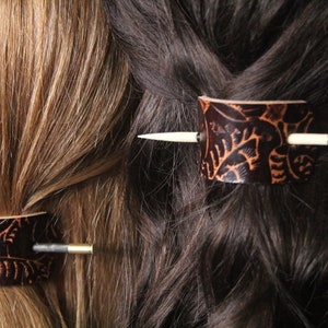 Puede incluir: Dos horquillas de cuero marr&oacute;n oscuro con dise&ntilde;os de hojas se insertan en cabello rubio y casta&ntilde;o oscuro. Cada horquilla tiene una pieza cil&iacute;ndrica de cuero y un alfiler de madera o metal.