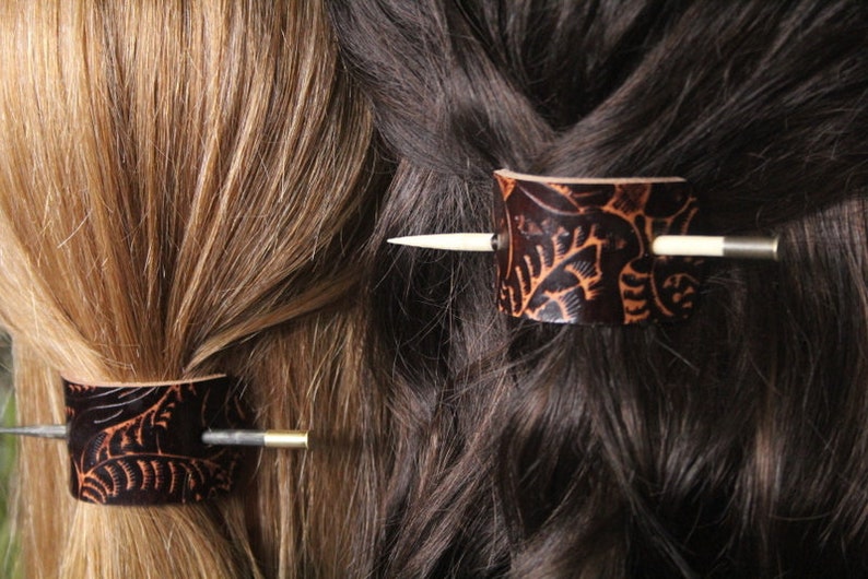 Puede incluir: Dos horquillas de cuero marr&oacute;n oscuro con dise&ntilde;os de hojas se insertan en cabello rubio y casta&ntilde;o oscuro. Cada horquilla tiene una pieza cil&iacute;ndrica de cuero y un alfiler de madera o metal.