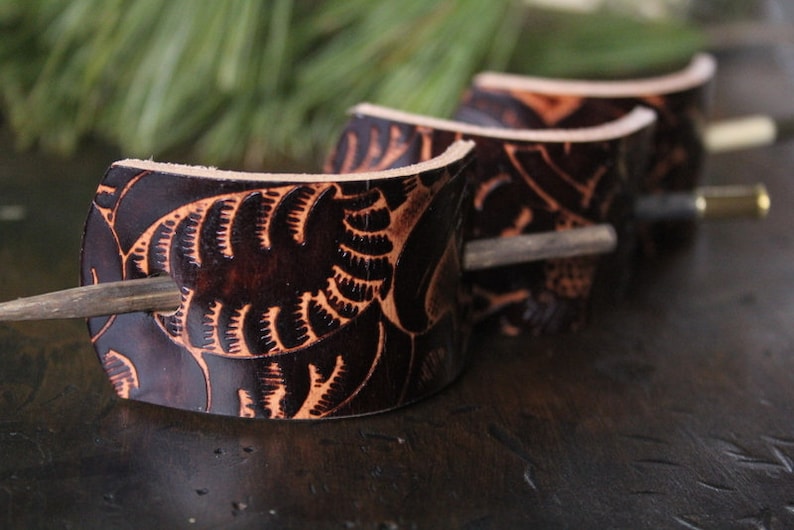 Puede incluir: Horquillas para el pelo de cuero marr&oacute;n oscuro con dise&ntilde;os florales tallados. Cada horquilla tiene una varilla de madera para asegurar el cabello. Las horquillas est&aacute;n dispuestas en fila, mostrando los intrincados detalles.