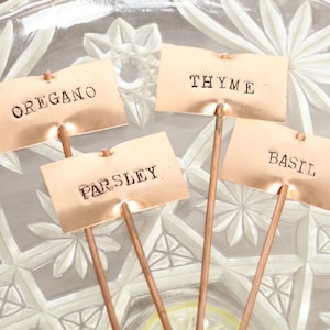 May include: Four copper plant markers with the words "Oregano", "Parsley", "Thyme", and "Basil" engraved on them.