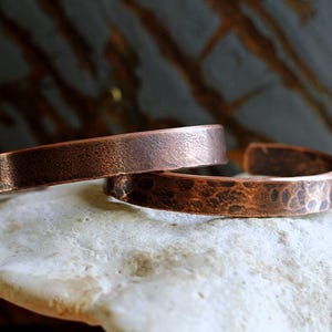 May include: Two copper cuff bracelets. One bracelet has a smooth, textured surface, while the other has a hammered, dimpled texture. The bracelets are a warm, reddish-brown color and are displayed on a light-colored, textured surface.