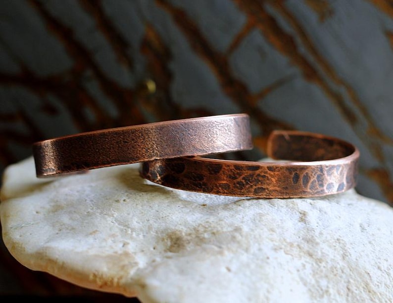 May include: Two copper cuff bracelets. One bracelet has a smooth, textured surface, while the other has a hammered, dimpled texture. The bracelets are a warm, reddish-brown color and are displayed on a light-colored, textured surface.