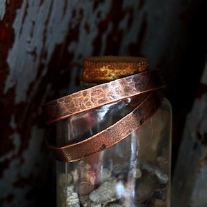May include: A clear glass jar filled with small stones, topped with a copper lid and wrapped with two hammered copper bands. The jar is set against a rustic, weathered wooden background.