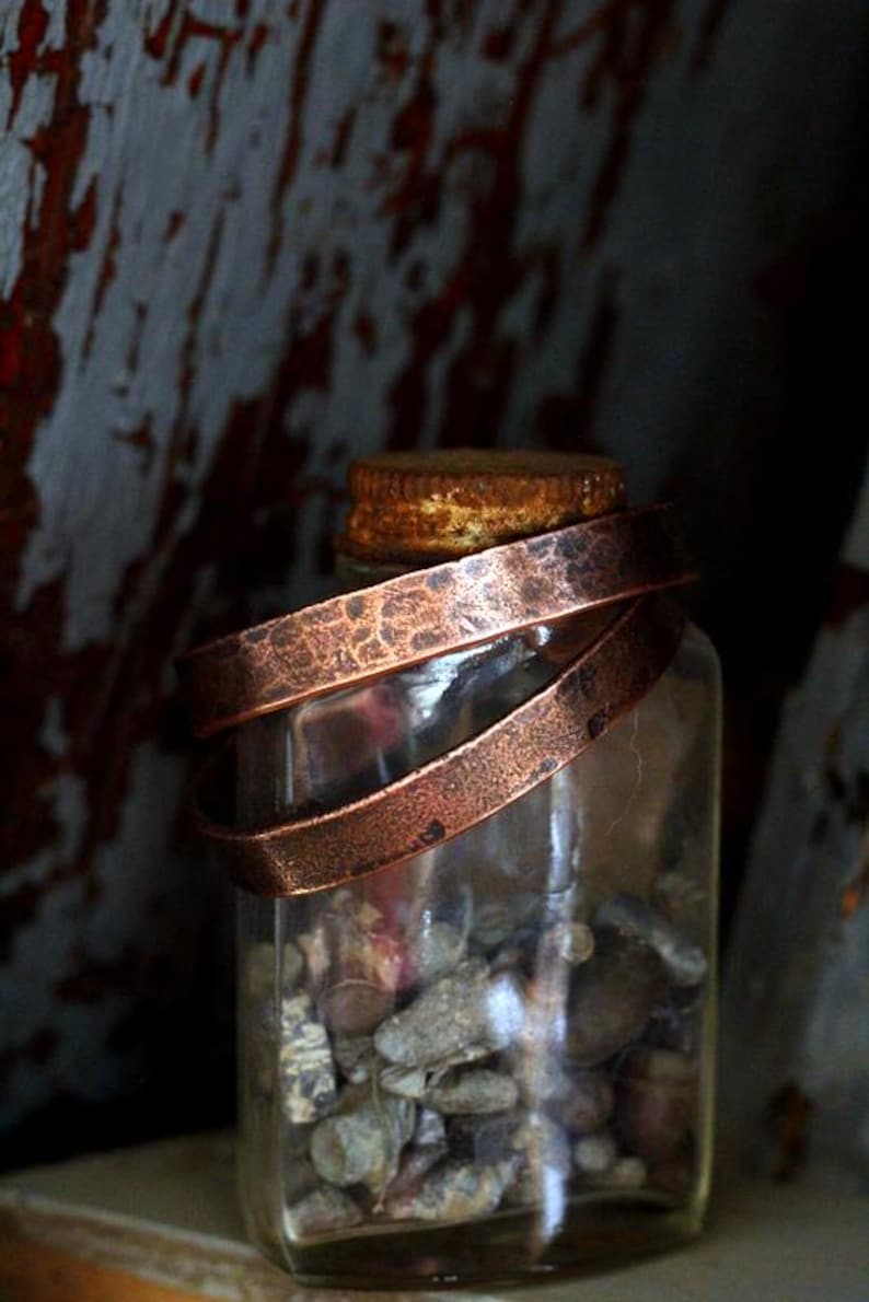 May include: A clear glass jar filled with small stones, topped with a copper lid and wrapped with two hammered copper bands. The jar is set against a rustic, weathered wooden background.