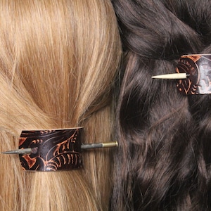 Puede incluir: Se muestran dos accesorios para el cabello, cada uno con una envoltura de cuero marr&oacute;n oscuro con un dise&ntilde;o de hojas en relieve. Un alfiler de metal asegura cada envoltura, sujetando el cabello en su lugar. Uno est&aacute; en cabello rubio, el otro en cabello casta&ntilde;o oscuro.