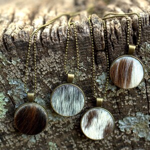 May include: Four necklaces with round pendants featuring a cowhide pattern. The pendants are brown, white, and black. The necklaces are on a wooden surface.