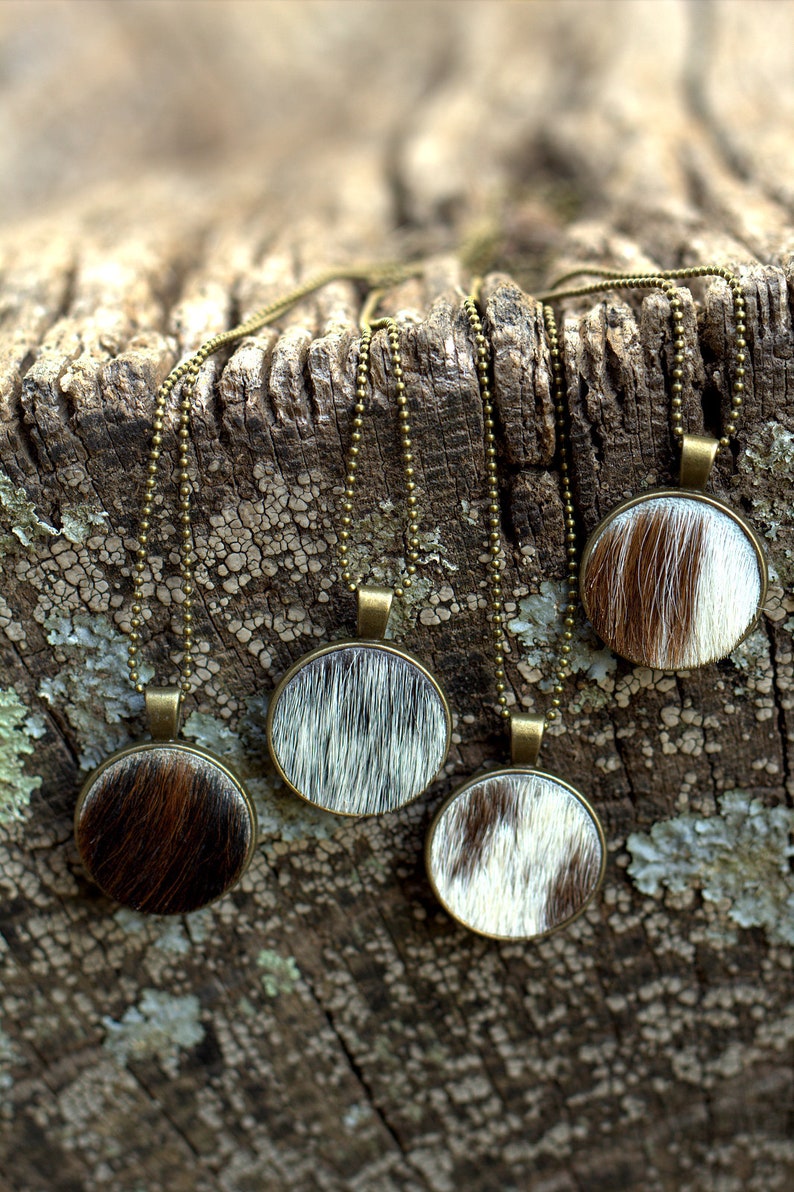 May include: Four necklaces with round pendants featuring a cowhide pattern. The pendants are brown, white, and black. The necklaces are on a wooden surface.