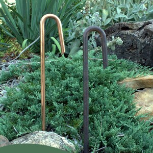 May include: Two metal garden stakes, one copper and one black, are shown in a garden setting. The copper stake has the text "Keep them polished (above left)" and the black stake has the text "Let them Age Gracefully (above right)".