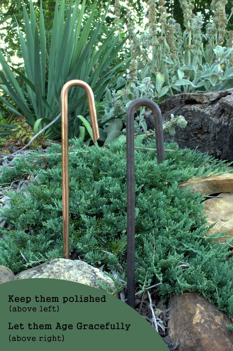 May include: Two metal garden stakes, one copper and one black, are shown in a garden setting. The copper stake has the text "Keep them polished (above left)" and the black stake has the text "Let them Age Gracefully (above right)".