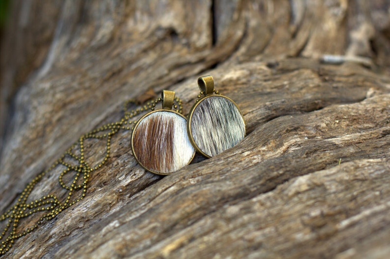 May include: Two round pendants with a brown and white cowhide pattern on a gold chain necklace. The necklace is lying on a piece of weathered wood.