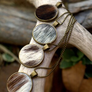 May include: Four round pendants with a brown and white cowhide pattern hang from a gold chain. The pendants are layered on top of each other.