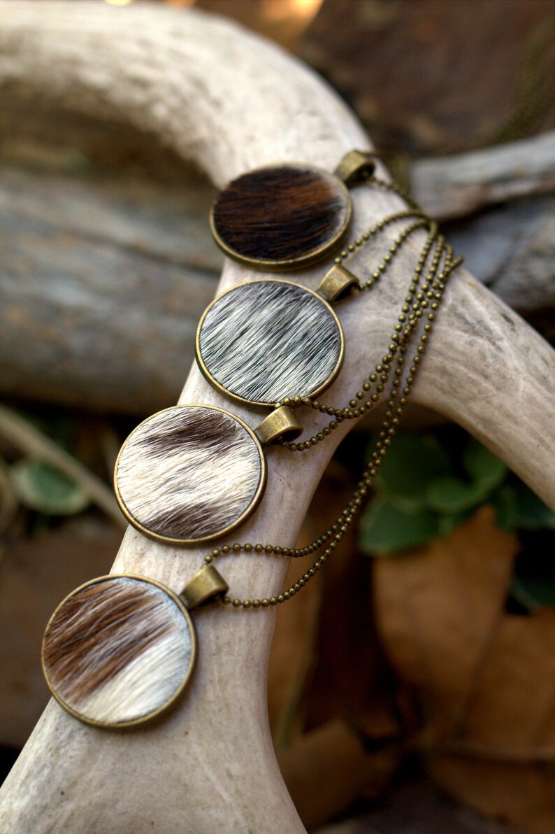 May include: Four round pendants with a brown and white cowhide pattern hang from a gold chain. The pendants are layered on top of each other.