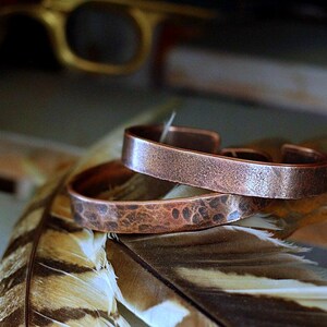 May include: Two copper cuff bracelets with textured surfaces are stacked on a feather. The bracelets have an open design and a hammered appearance. The background includes a blurred gold-colored object.