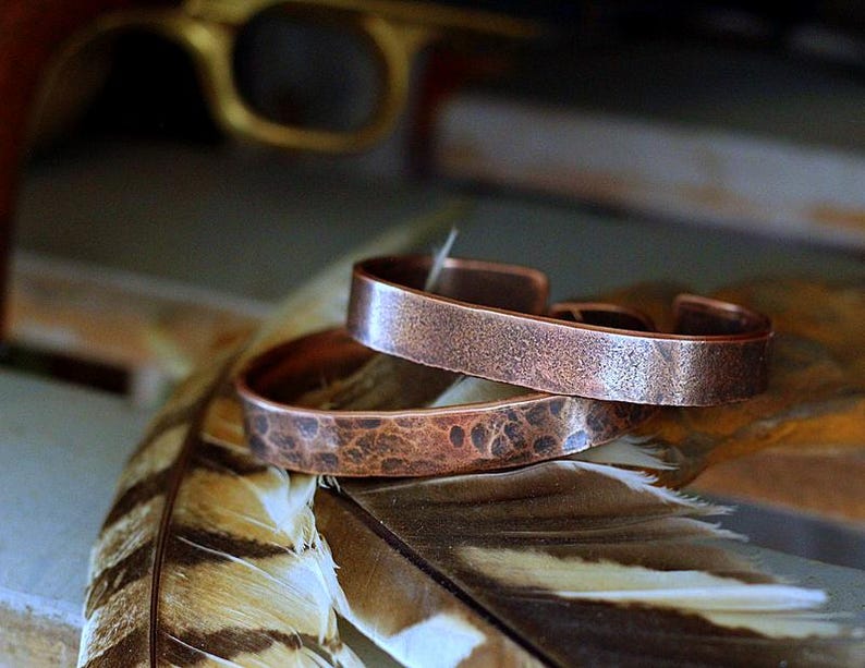 May include: Two copper cuff bracelets with textured surfaces are stacked on a feather. The bracelets have an open design and a hammered appearance. The background includes a blurred gold-colored object.