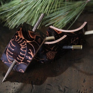 Puede incluir: Dos horquillas para el pelo de cuero marr&oacute;n oscuro con dise&ntilde;os florales tallados. Cada horquilla tiene un pasador de madera y una pieza decorativa de cuero. El cuero es de color marr&oacute;n intenso con acentos marrones claros. El fondo incluye agujas de pino.
