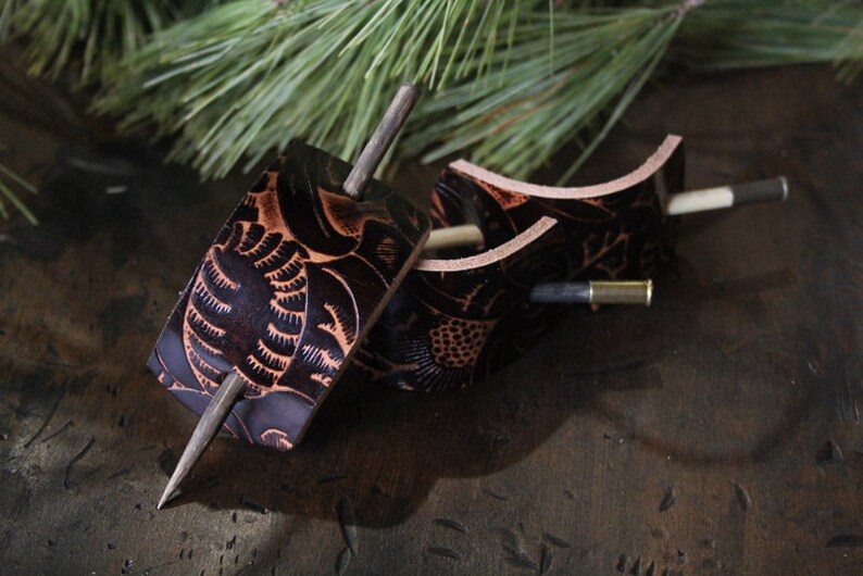 Puede incluir: Dos horquillas para el pelo de cuero marr&oacute;n oscuro con dise&ntilde;os florales tallados. Cada horquilla tiene un pasador de madera y una pieza decorativa de cuero. El cuero es de color marr&oacute;n intenso con acentos marrones claros. El fondo incluye agujas de pino.