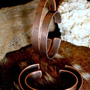 May include: Three copper cuff bracelets with a rustic, hammered texture. The open-ended design allows for easy wear. The bracelets are displayed on a brown and tan fur-like surface, with a blurred background.