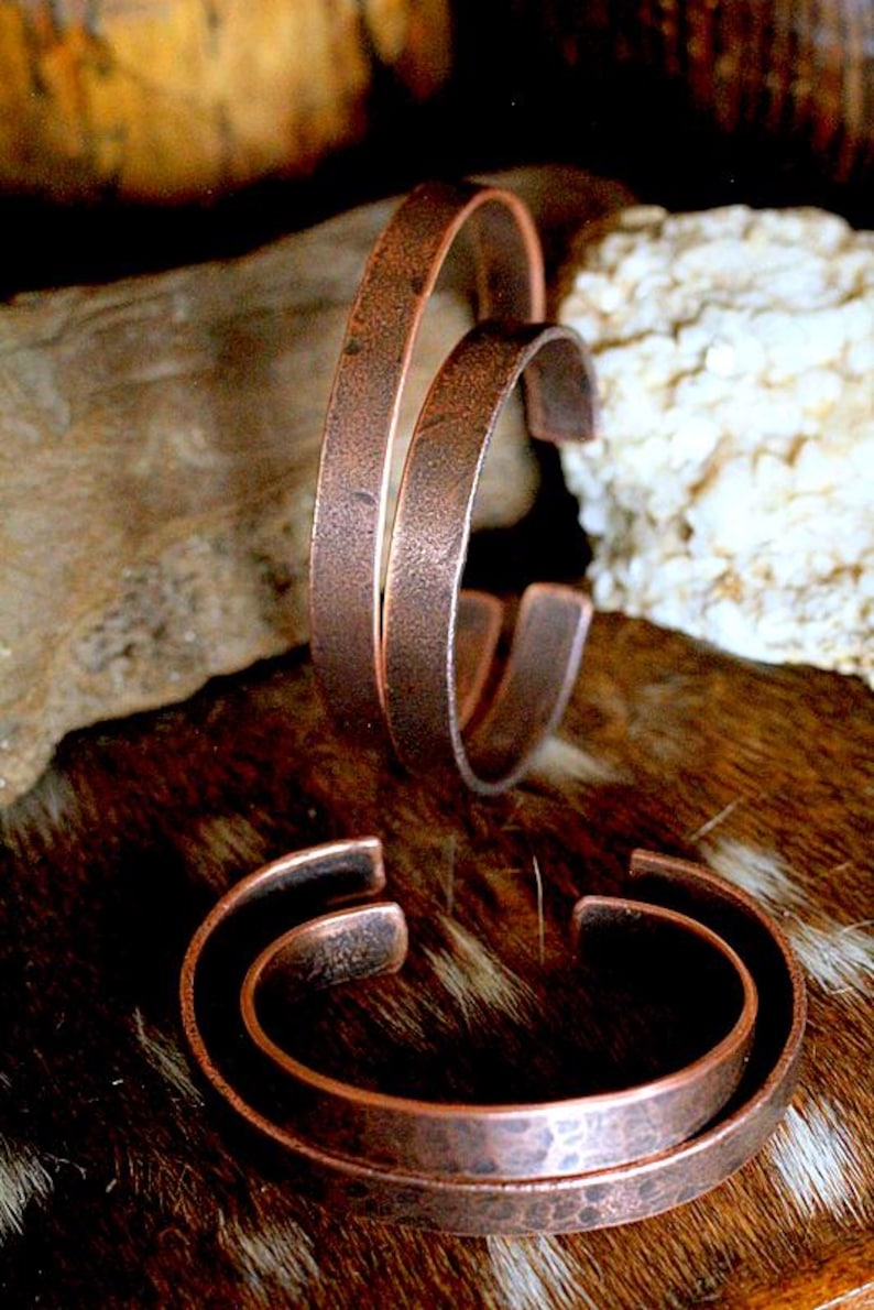 May include: Three copper cuff bracelets with a rustic, hammered texture. The open-ended design allows for easy wear. The bracelets are displayed on a brown and tan fur-like surface, with a blurred background.
