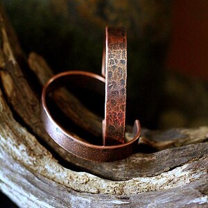 May include: Two copper-colored cuff bracelets with a textured finish. One bracelet is standing upright, while the other is lying down. The bracelets are displayed on a piece of weathered wood, showcasing their rustic appeal.